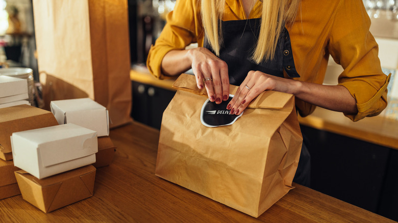 Woman wearing a yellow shirt and black apron packaging to-go foods in a bag at restaurant