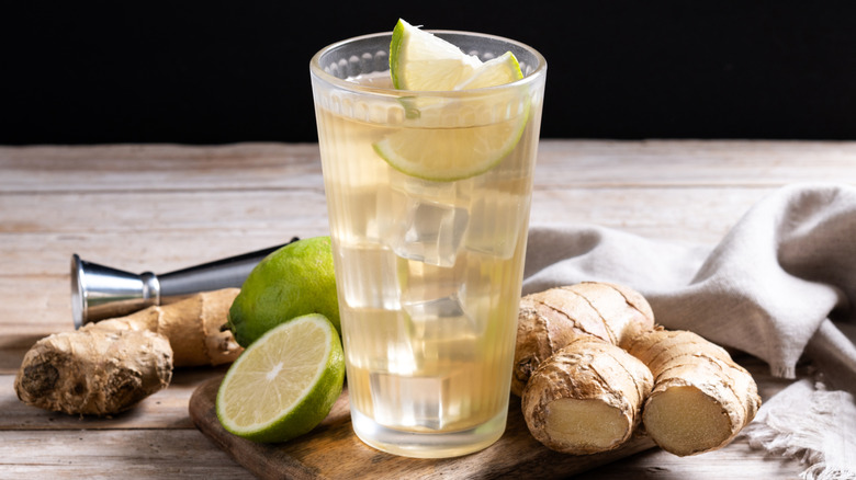 Glass of ginger ale over ice surrounded by sliced ginger and limes.
