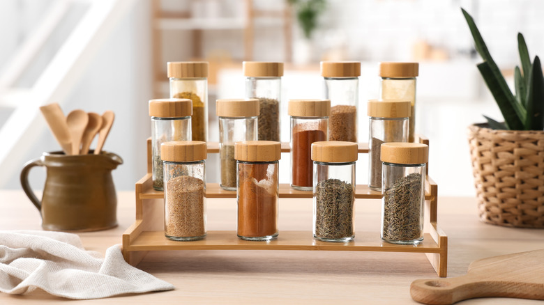 A selection of 12 spices on three raised platforms with a plant and a pot of cooking utensils next to them