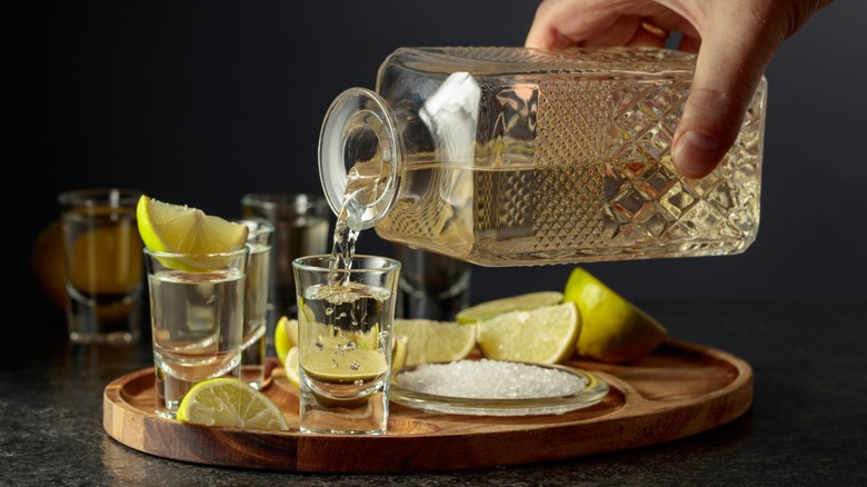 A person pouring tequila into a glass on a wooden tray with salt and limes