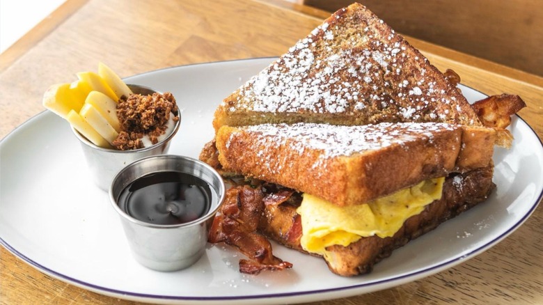 Eggs and bacon on a french toast sandwich on white plate with sides of sauce