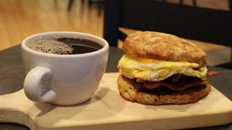 Biscuit sandwich with bacon, eggs, and cheese on a wooden board with black coffee