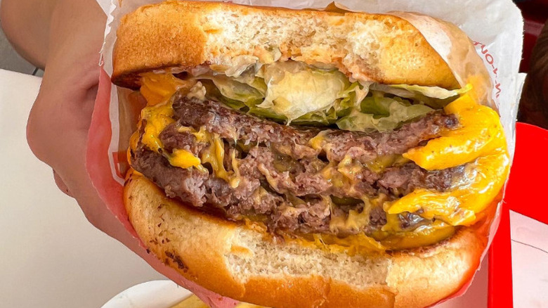 a person holds an In-N-Out burger with three patties and three slices of cheese and one bite taken out up to the camera.