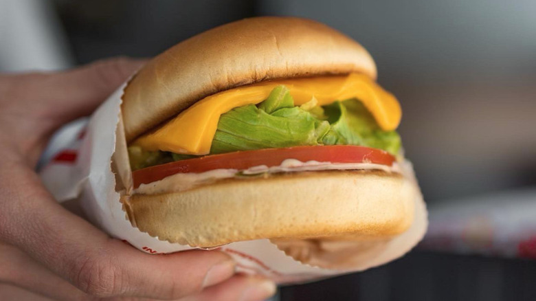 a hand holds a wrapped grilled cheese on a burger bun with lettuce, tomato, and spread.