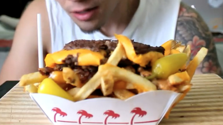 a basket of fries with two burger patties, melted cheese, banana peppers, grilled onions, and spread set on a wooden board in front of a man.