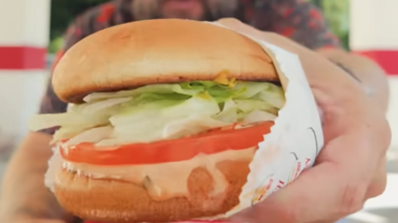 a man's hand holds a burger with lettuce, tomato, cheese, onions, and spread, but no meat.