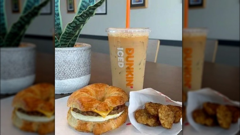 A sausage, egg, and cheese croissant, hashbrowns, and an iced coffee from Dunkin' on a table in a home next to a potted plant