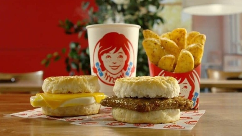 Two Wendy's biscuit breakfast sandwiches, seasoned potatoes, and a drink on a table in a living room