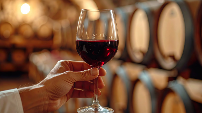 A hand holding a glass of red wine in front of rows of barrels