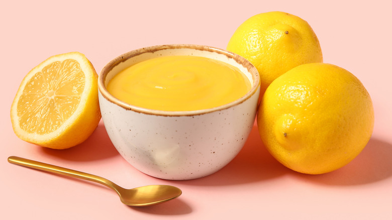 A bowl of lemon curd on a pink table with lemons and a spoon beside it