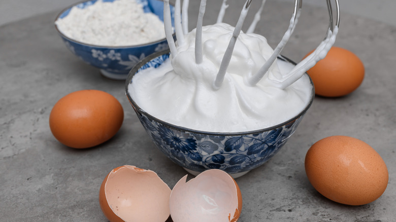 Meringue with a whisk in a bowl, with eggs and a bowl of powdered sugar on the side