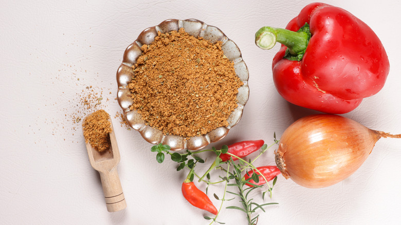 Cajun seasoning in a bowl and a wooden spoon with peppers and an onion on the side, all on a white table