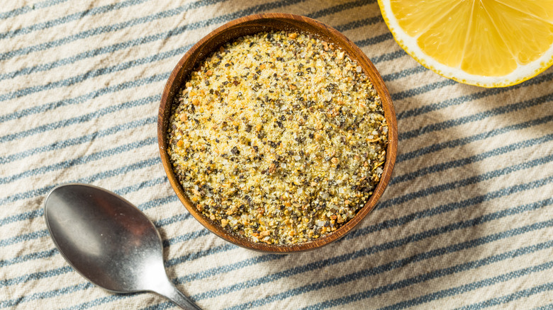 Lemon pepper seasoning in a wooden bowl placed on a table with half a lemon and a spoon on the side