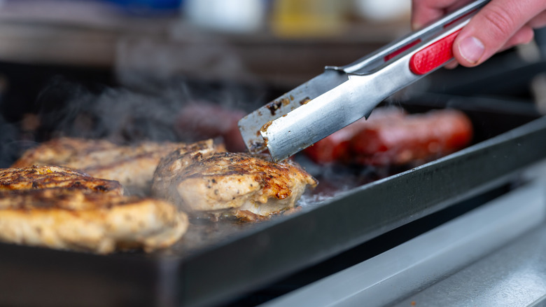 A hand holding tongs and flipping chicken breasts on a griddle.