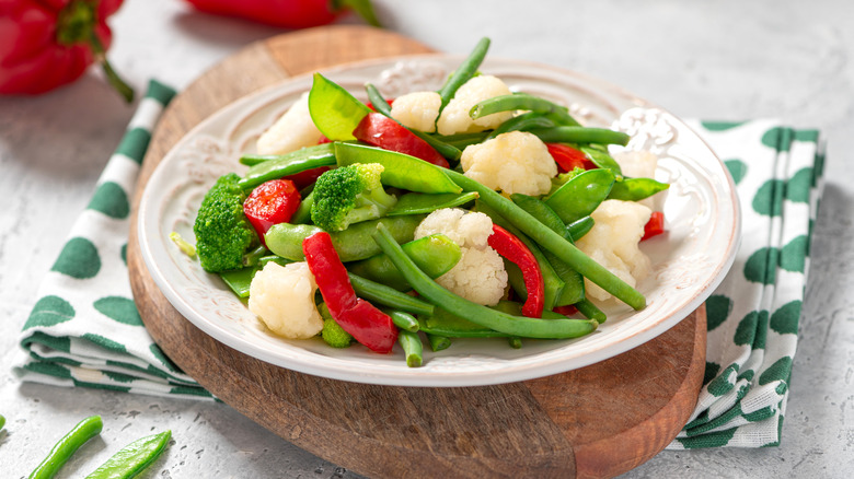 Steamed vegetables including cauliflower, red peppers, broccoli, green beans, and pea pods on a white plate