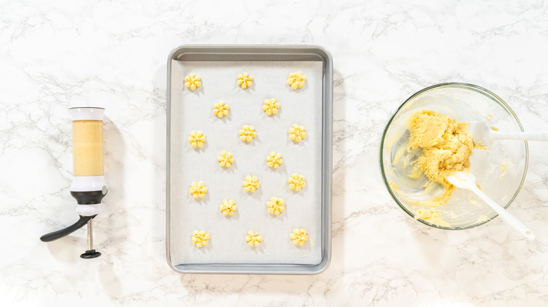 Cookie press, baking pan with shaped cookie dough, and bowl of dough on marble surface