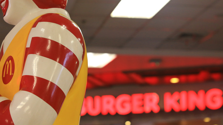 A red neon Burger King logo in the background, peeking out from behind the shoulder of a Ronald McDonald statue in the foreground.