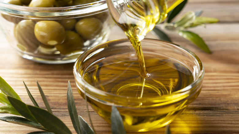Golden olive oil poured into a small clear bowl with olives in background