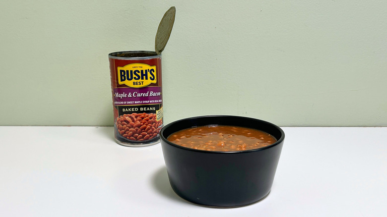 A black bowl of baked beans next to an open can of Bush's Best Maple and Cured Bacon Baked Beans