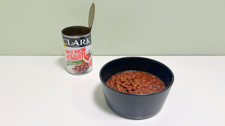 A black bowl of baked beans next to an open can of Clark Apple Bacon Mesquite Gourmet Baked Beans
