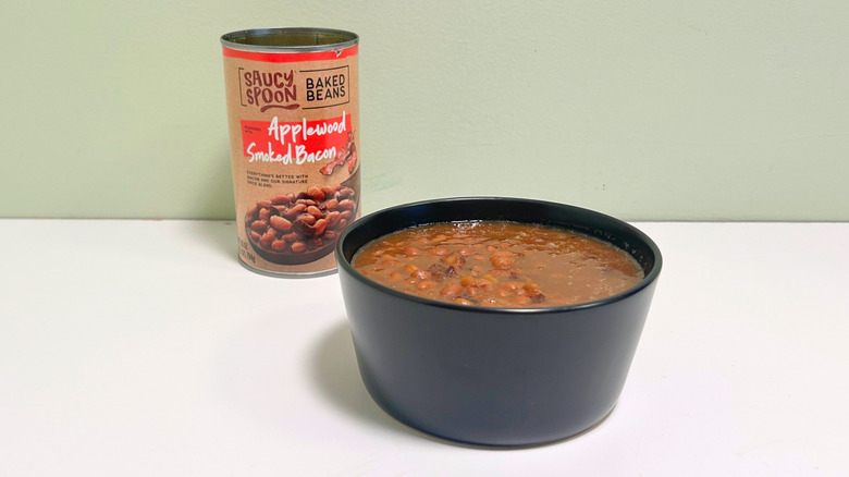 A black bowl of baked beans next to an open can of Saucy Spoon Applewood Smoked Bacon Baked Beans