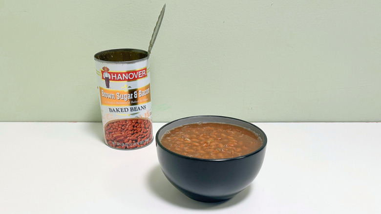 A black bowl of baked beans next to an open can of Hanover Brown Sugar and Bacon Baked Beans