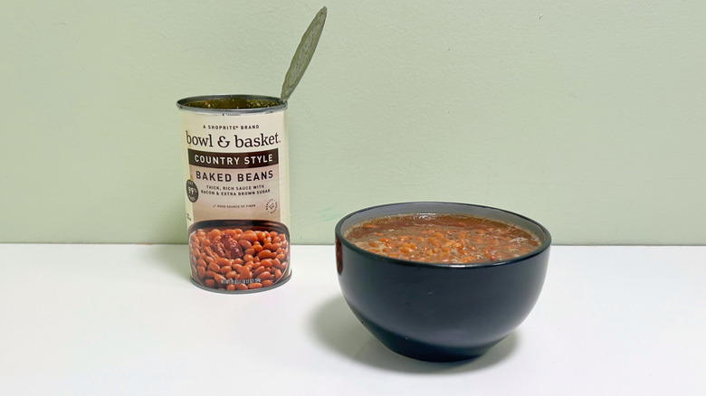 A black bowl of baked beans next to an open can of Bowl and Basket Country Style Baked Beans