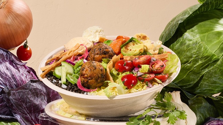 A bowl of Cava food with meatballs and sliced vegetables surrounded by fresh produce