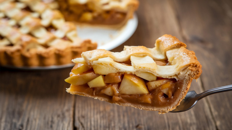 A fork holds a slice of apple pie, with an out-of-focus pie in the background