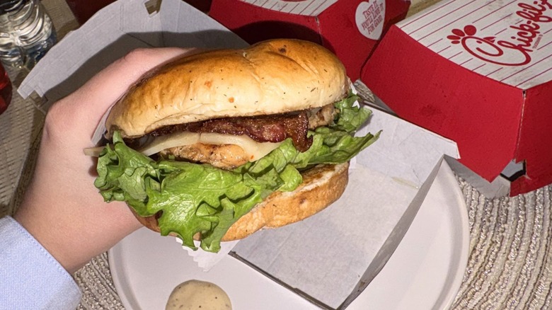 Hand holding up a grilled Jalapeño Ranch Club Chicken Sandwich with sauce on the side of a plate underneath