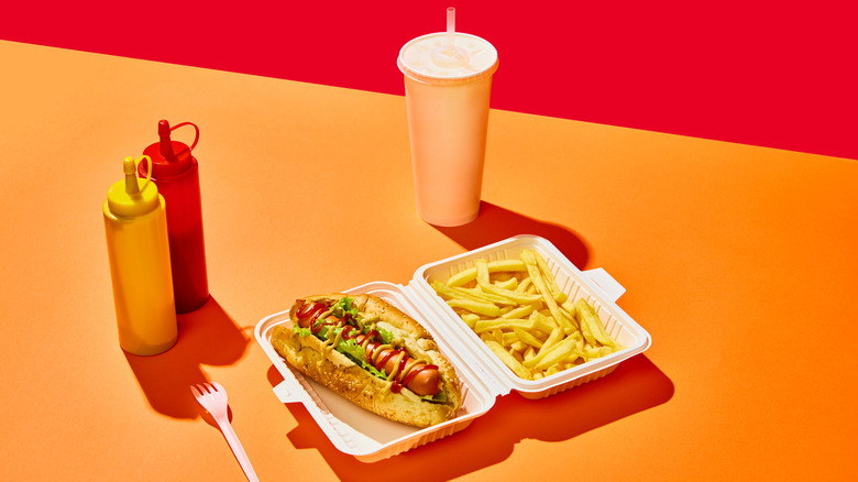 A hot dog and fries sit in a food carton next to a drink and condiments, against an orange and red background
