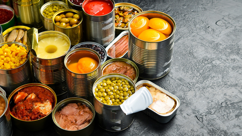 An assortment of open cans of food including peaches, beans, corn, pineapple, and fish on a slate grey background
