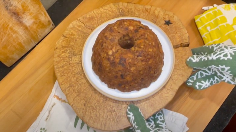A pair of gloved hands hold a board, upon which sits a steamed carrot pudding