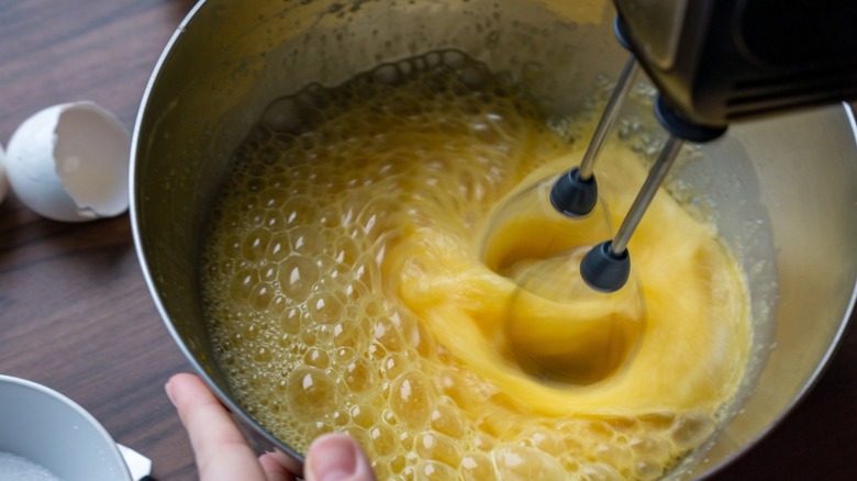 A person whipping eggs with sugar in a bowl using a hand mixer