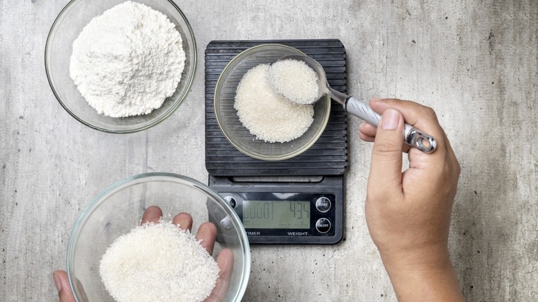 A person weighing out sugar in a bowl on a kitchen scale with a bowl of flour on the side