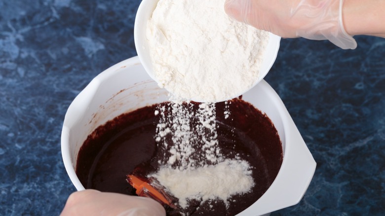 A person adding flour to a white bowl full of brownie batter