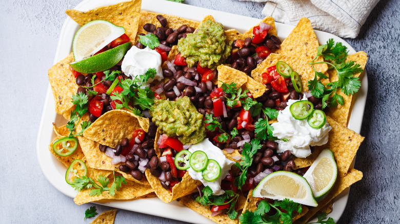 A tray of vegan nachos with black beans, avocado, sour cream, cilantro, lime, and tomatoes.