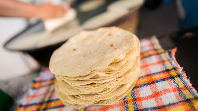 Fresh tortillas piled on a towel, as someone heats more tortillas on a large comal in the background
