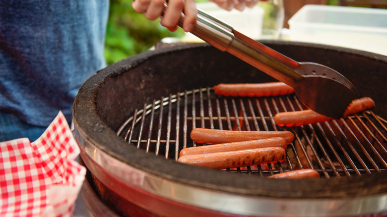 A hand with tongs turning hot dogs on a round charcoal grill
