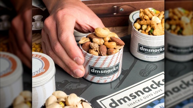 a hand holding an open tin of mixed nuts flanked by another open tin