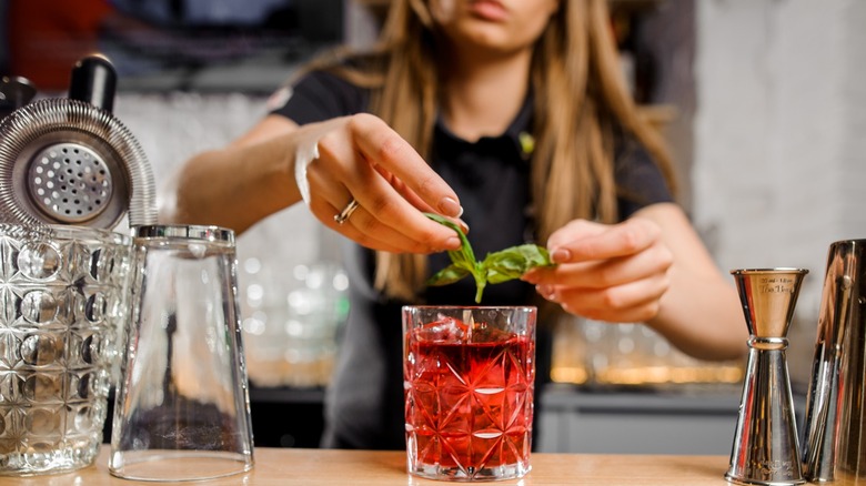 woman adding garnish to a red cocktail with glassware and jiggers on the side