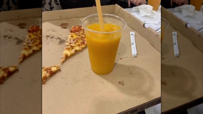 A Costco mango smoothie sits in a pizza box with a leftover slice of pizza next to it