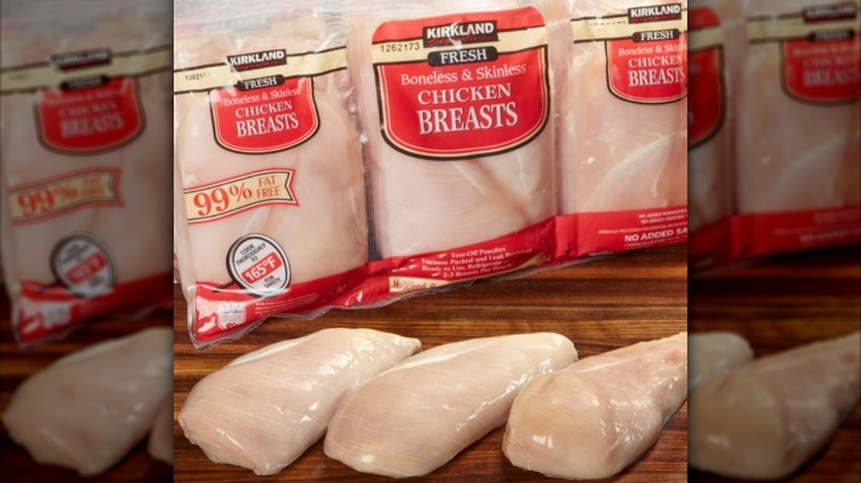 Three packages of Kirkland Signature boneless and skinless chicken breasts and three raw chicken breasts on a wooden board