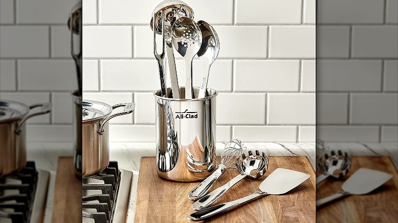 a container with All-Clad utensils on a wooden board with 3 utensils on it, placed next to the kitchen stove