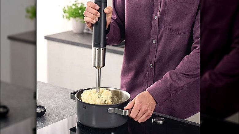 man in a maroon shirt holding handheld blender in a pot