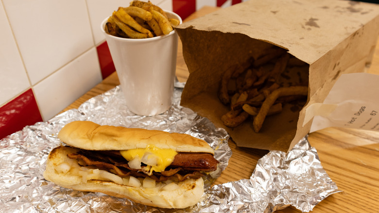 A grilled hot dog from Five Guys, topped with cheese, bacon, and onions, along with a side of french fries and several more fries in the bag.