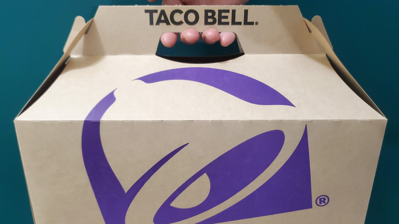 A hand holding a cardboard Taco Bell takeout box with a purple logo against a dark teal background.