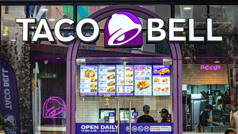 Glass wall exterior of a Taco Bell restaurant with the purple and white bell logo alongside white "Taco Bell" text and the menu inside.