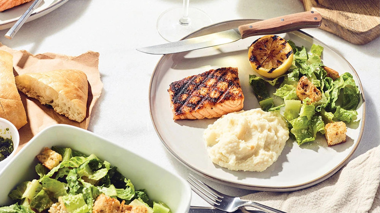 a table with a plate of grilled salmon, side Caesar salad, and side mashed potatoes, with a basket of bread and extra salad visible