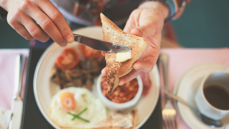 A close-up of hands buttering a piece of toast, with a plate of breakfast underneath